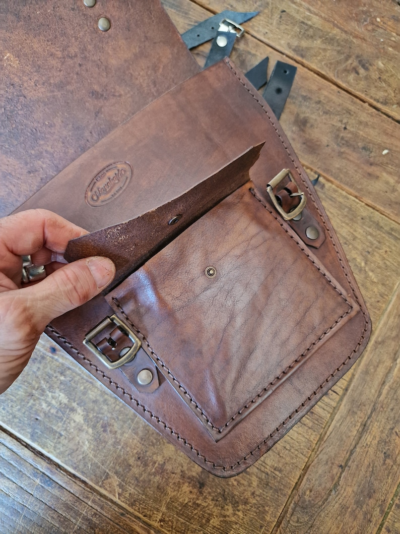 May include: A brown leather motorcycle saddlebag with a flap that opens to reveal a smaller pocket. The bag has a brass buckle and stitching around the edges. The bag has a leather label that says "Wayward" with a small logo.