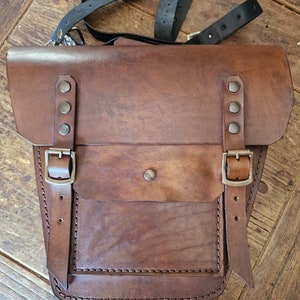 May include: A brown leather motorcycle saddlebag with a flap and two buckles. The bag has a smaller pocket on the front and is stitched with brown thread.