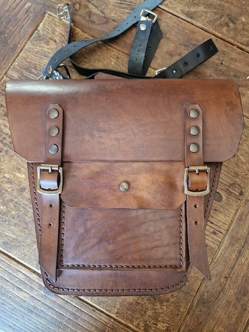 May include: A brown leather motorcycle saddlebag with a flap and two buckles. The bag has a smaller pocket on the front and is stitched with brown thread.