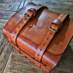 Può includere: Una borsa in pelle marrone con due cinturini regolabili e fibbie in ottone. La borsa ha una forma rettangolare ed è realizzata in pelle di alta qualità.