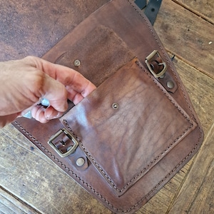 May include: A brown leather motorcycle saddlebag with a flap and a buckle closure. The bag has a smaller pocket on the front with a buckle closure.