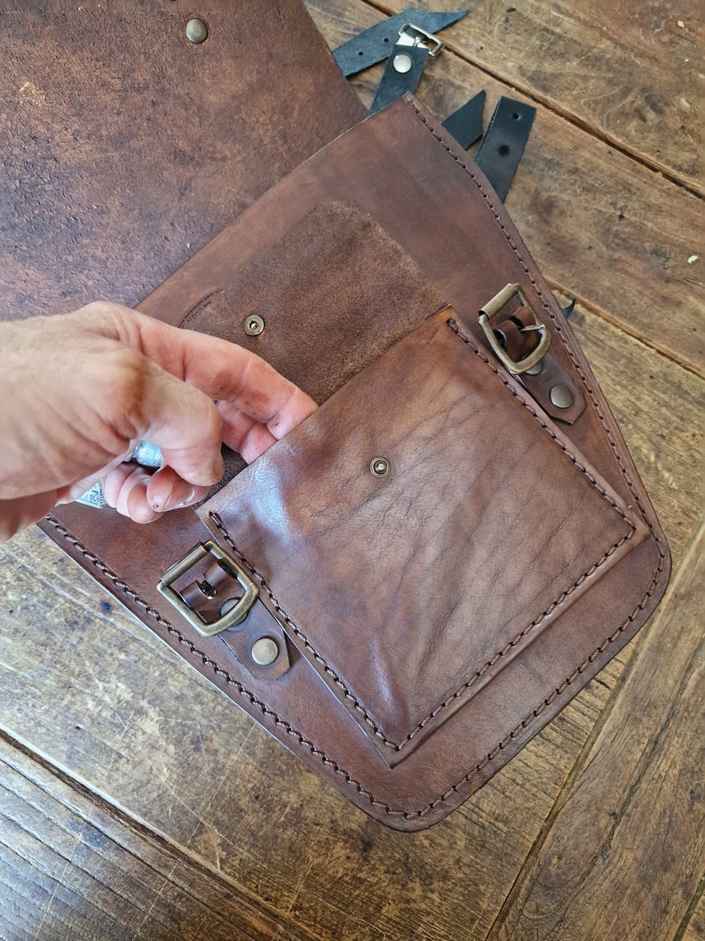 May include: A brown leather motorcycle saddlebag with a flap and a buckle closure. The bag has a smaller pocket on the front with a buckle closure.