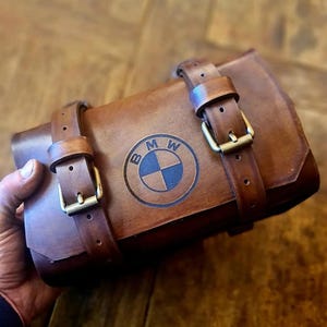 Può includere: Un rotolo porta attrezzi per moto in pelle marrone con due cinturini e fibbie. Il rotolo ha il logo BMW in rilievo sul davanti.