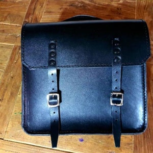 Può includere: Una borsa a tracolla in pelle nera con due cinturini regolabili e fibbie argentate. La borsa ha una chiusura a lembo e un design cucito.