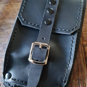 May include: Black leather pouch with a silver buckle and three black snaps. The pouch has a flap with a stitched edge.