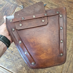 May include: A brown leather pouch with a flap and multiple silver rivets. The pouch has a stitched design and is secured with a strap.