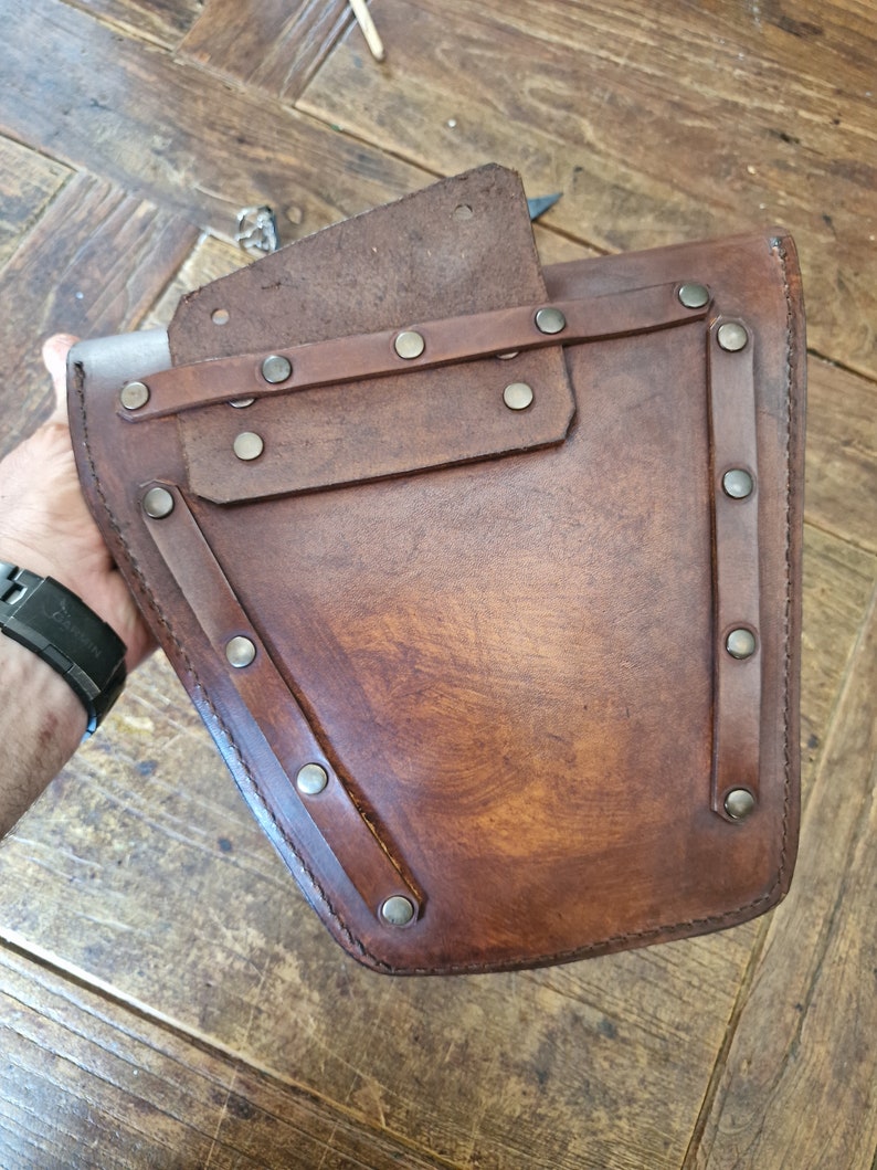 May include: A brown leather pouch with a flap and multiple silver rivets. The pouch has a stitched design and is secured with a strap.