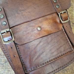 May include: A brown leather satchel bag with a flap closure and a front pocket. The bag has a strap and metal buckles.