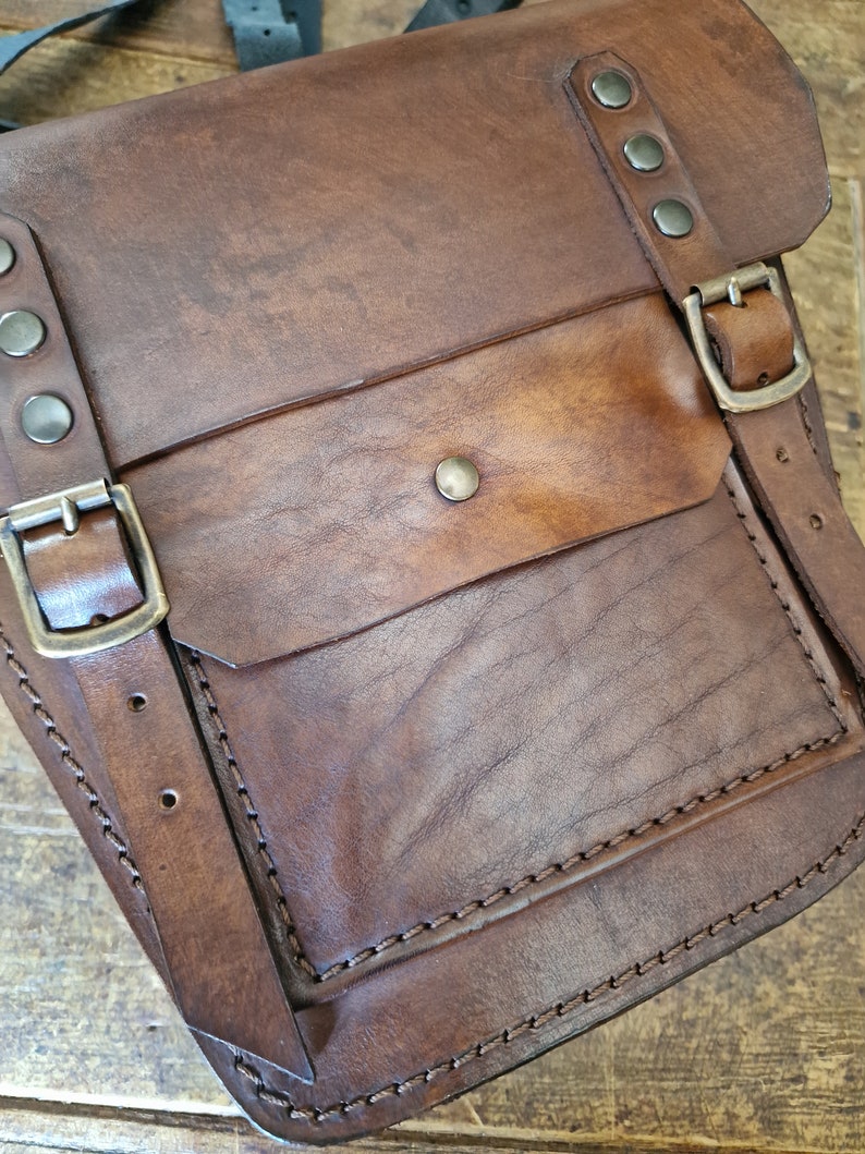 May include: A brown leather satchel bag with a flap closure and a front pocket. The bag has a strap and metal buckles.
