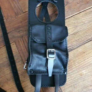 May include: Black leather motorcycle tank bag with a large pocket and adjustable straps. The bag has a circular opening and silver buckles.