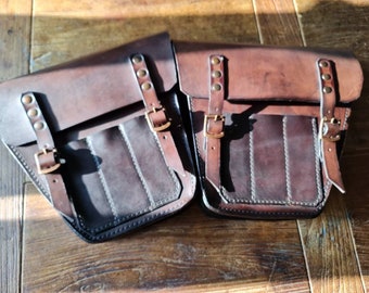 May include: Two brown leather motorcycle saddlebags with buckles and straps. The bags have a dark brown, almost black, color and are made of a textured leather.