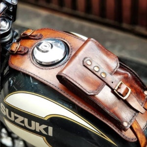 Puede incluir: Una bolsa de depósito de motocicleta de cuero marrón con cierre de hebilla y un pequeño bolsillo en la parte delantera. La bolsa está unida a un depósito de gasolina de motocicleta negro con la palabra "SUZUKI" en letras blancas.