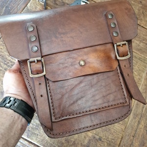 May include: A brown leather satchel bag with a flap closure, two buckles, and a large front pocket. The bag has a distressed look and is stitched with brown thread.