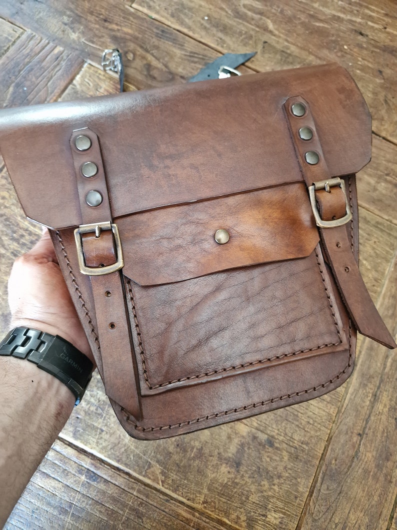 May include: A brown leather satchel bag with a flap closure, two buckles, and a large front pocket. The bag has a distressed look and is stitched with brown thread.