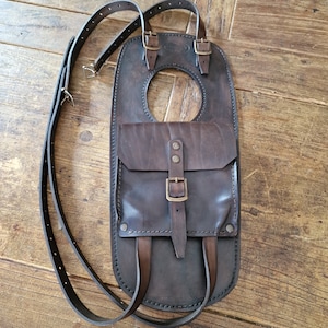 Puede incluir: Una bolsa de depósito de motocicleta de cuero marrón con cierre de solapa y un pequeño bolsillo frontal. La bolsa tiene una correa que va sobre el hombro y otra correa que va alrededor de la cintura.