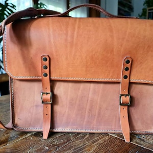 May include: A brown leather satchel bag with two adjustable straps and brass buckles. The bag has a flap closure and a white stitching detail.