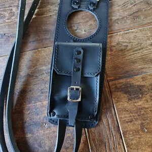 May include: Black leather motorcycle tank bag with a small pocket and two straps. The bag has a buckle closure and a circular opening.