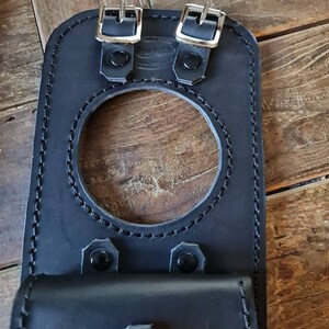 May include: Black leather motorcycle gas tank bag with two buckles and a flap. The bag has a circular opening for the gas cap.
