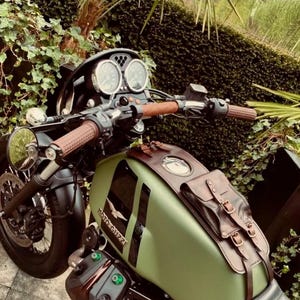 Może przedstawiać: Zielony motocykl z brązowymi skórzanymi sakwami i kierownicą. Motocykl ma okrągłą lampę przednią i cyfrowy prędkościomierz. Motocykl jest zaparkowany przed zielonym krzewem.