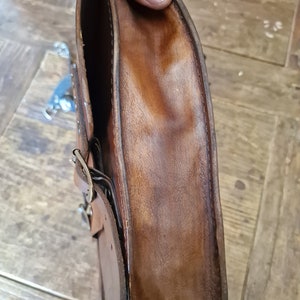 May include: A brown leather motorcycle saddlebag with a metal buckle and stitching. The bag is folded open to show the inside lining.