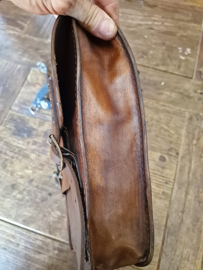 May include: A brown leather motorcycle saddlebag with a metal buckle and stitching. The bag is folded open to show the inside lining.