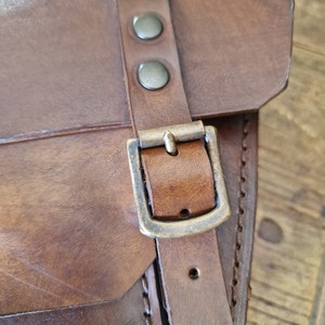 May include: A brown leather bag with a brass buckle and rivets. The bag has a flap with a stitched edge.