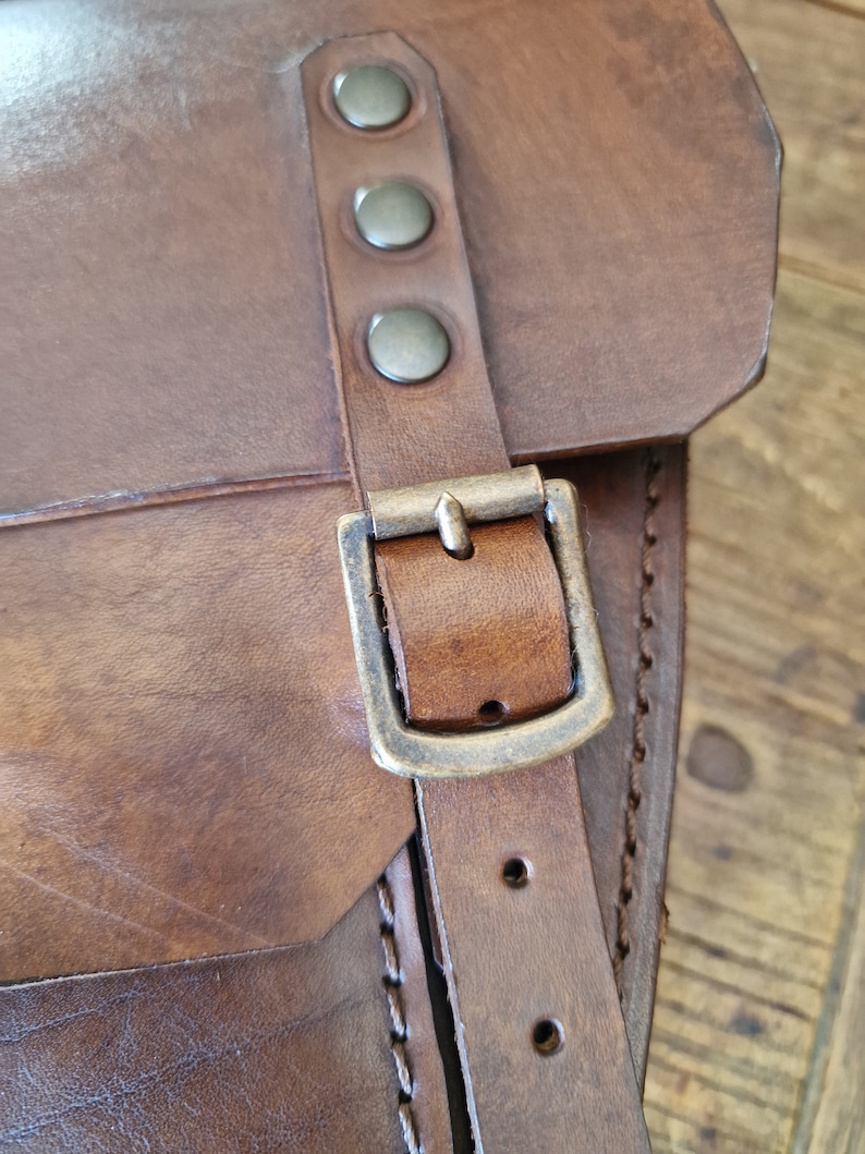 May include: A brown leather bag with a brass buckle and rivets. The bag has a flap with a stitched edge.