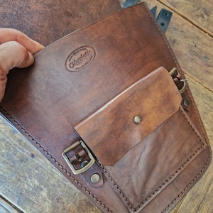 May include: Brown leather motorcycle saddlebag with a flap and a smaller pocket. The bag has a brass buckle and stitching detail. The bag has a leather label with the text "Maxwells Est. 1995".