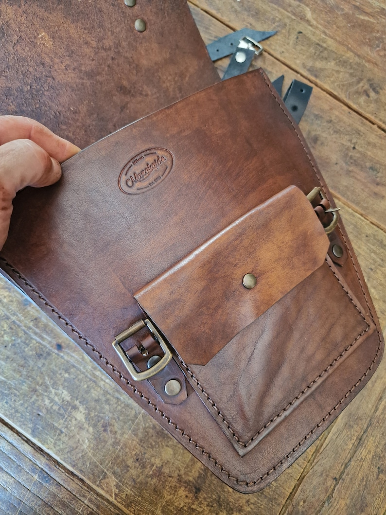 May include: Brown leather motorcycle saddlebag with a flap and a smaller pocket. The bag has a brass buckle and stitching detail. The bag has a leather label with the text "Maxwells Est. 1995".