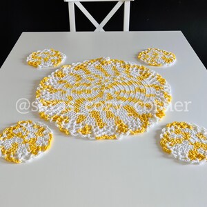 May include: A white table setting featuring a large, round, crocheted placemat and four matching coasters. The placemat and coasters have a spiral design with yellow and white colors. The placemat is approximately 12 inches in diameter.