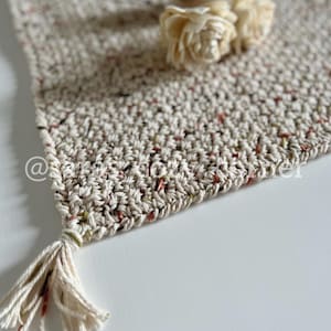 May include: A beige crocheted table runner with a fringe tassel. The runner has a speckled pattern with red, green, and brown flecks. Two white dried flowers are on top of the runner.