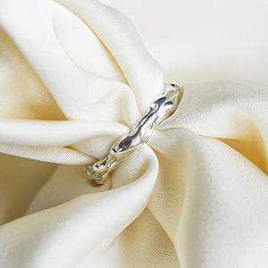 Può includere: Un anello d'argento con un design testurizzato e organico. L'anello è mostrato su uno sfondo di tessuto bianco.
