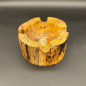 May include: A handcrafted ashtray made from wood, featuring a natural, textured appearance with shades of yellow, brown, and black. The ashtray has a circular bowl shape with a coin in the center and two grooves for resting cigarettes.