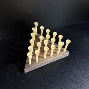Pode incluir: Um suporte triangular de madeira para tees de golfe. A base de madeira castanha escura contém inúmeros tees de golfe bege, dispostos em uma grade. Os tees de golfe são apresentados de forma organizada.