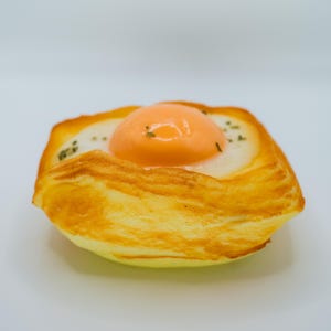 May include: A golden brown pastry with a flaky crust filled with a bright orange egg yolk and a white egg white. The pastry is topped with green herbs.