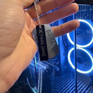 Puede incluir: Una placa de identificación plateada con el texto "(head, jaws)" y una cadena. La placa es rectangular con esquinas redondeadas y un borde negro. El fondo es una caja de ordenador con luces azules.
