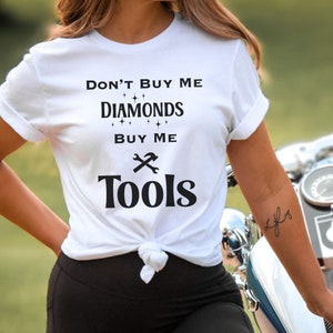 Puede incluir: Camiseta blanca con texto negro que dice "No me compres diamantes, c&oacute;mprame herramientas". El texto est&aacute; rodeado de peque&ntilde;as estrellas. Una llave inglesa y un martillo est&aacute;n representados sobre la palabra "herramientas".
