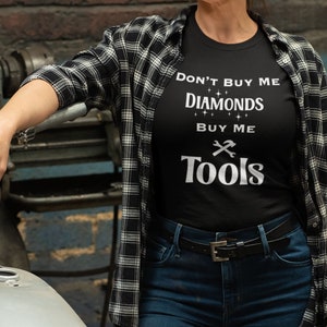 Puede incluir: Una camiseta negra con el texto "No me compres diamantes, c&oacute;mprame herramientas" en letras blancas. La camiseta la lleva puesta una persona con una camisa de cuadros y vaqueros.