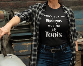 Puede incluir: Una camiseta negra con el texto "No me compres diamantes, cómprame herramientas" en letras blancas. La camiseta la lleva puesta una persona con una camisa de cuadros y vaqueros.