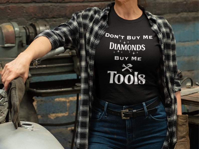 Puede incluir: Una camiseta negra con el texto "No me compres diamantes, c&oacute;mprame herramientas" en letras blancas. La camiseta la lleva puesta una persona con una camisa de cuadros y vaqueros.