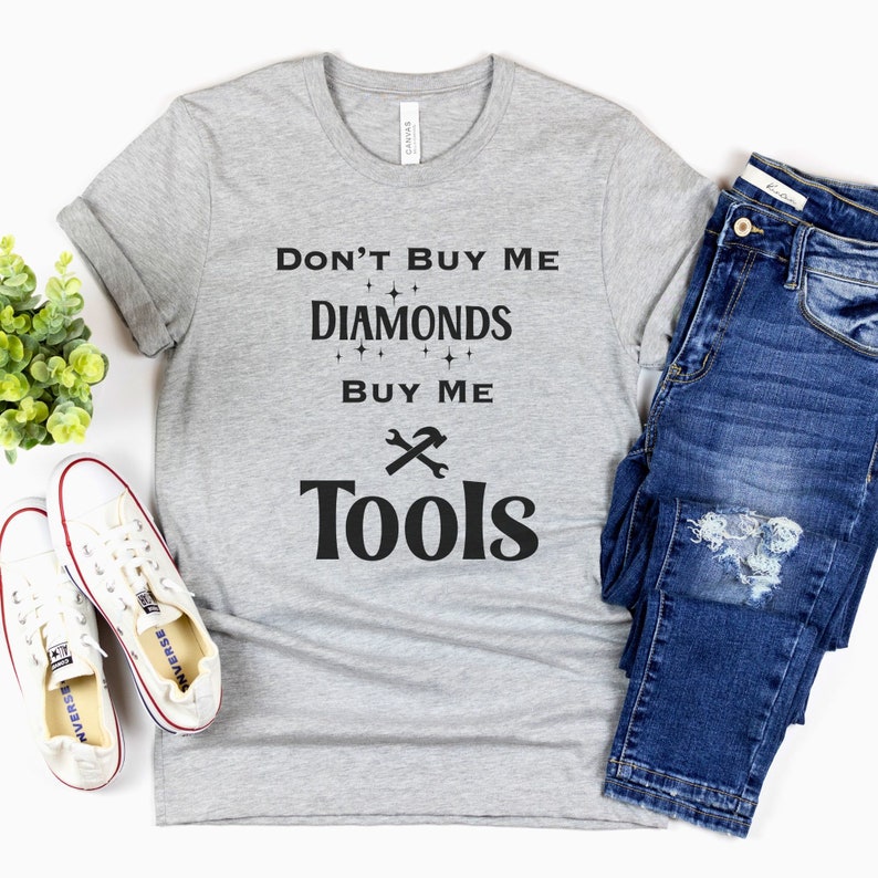 Puede incluir: Camiseta gris con texto negro que dice "No me compres diamantes, c&oacute;mprame herramientas". El texto est&aacute; rodeado de peque&ntilde;os diamantes. Una llave inglesa y un martillo est&aacute;n cruzados debajo del texto.