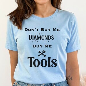 Puede incluir: Una camiseta azul claro con el texto "No me compres diamantes, c&oacute;mprame herramientas" en negro. El texto est&aacute; rodeado de peque&ntilde;as estrellas y un gr&aacute;fico de llave inglesa y martillo.