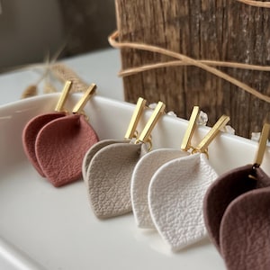 Op de afbeelding: Een verzameling blad-vormige oorbellen in diverse neutrale kleuren, waaronder terracotta, beige en wit. Elke oorbel heeft een getextureerd oppervlak en een goudkleurige rechthoekige bovenkant. De oorbellen worden gepresenteerd op een wit dienblad.