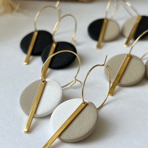 Könnte beinhalten: Goldene Creolen-Ohrringe mit geometrischem Design. Jeder Ohrring hat einen kreisförmigen, zweifarbigen Anhänger in Schwarz, Weiß und Beige, der von einem goldenen Balken geteilt wird. Die Ohrringe werden auf einer weißen Oberfläche präsentiert.