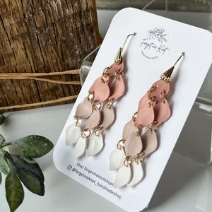 Puede incluir: Pendientes colgantes con un diseño de pétalos en capas en tonos rosa, beige y blanco. Los pendientes están sujetos a ganchos dorados y se exhiben en una tarjeta blanca con el texto "Forget me Knot Handmade Shop".