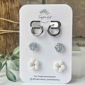 May include: A set of three pairs of earrings on a white display card labelled Forget me Knot. The top pair are silver-tone hoops, the middle pair are hexagonal studs with a light blue leaf pattern, and the bottom pair are white five-petal flower studs.
