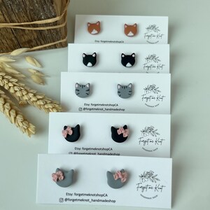Op de afbeelding: Stud oorbellen in katvorm in verschillende kleuren: oranje, zwart, grijs en grijs met roze strikjes. Elk paar wordt gepresenteerd op een witte kaart met de tekst "Forget Me Knot" en Etsy shop informatie.