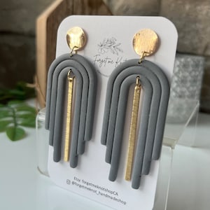 Peut inclure: Boucles d'oreilles en argile polymère grise avec des accents dorés. Les boucles d'oreilles sont en forme d'arc-en-ciel avec une barre dorée verticale au centre. Les boucles d'oreilles sont présentées sur une carte blanche avec le texte "Forget me Knot" et "Handmade".