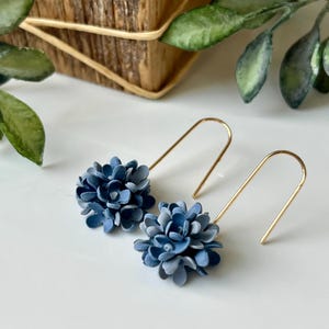 Peut inclure: Une paire de boucles d'oreilles composée de grappes de petites fleurs bleu mat fixées à de fins fils métalliques dorés en forme de U. Les éléments floraux sont densément superposés, créant un aspect arrondi et texturé sur chaque boucle.