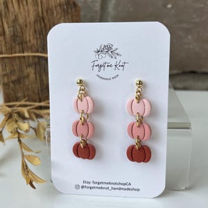 May include: Dangle earrings featuring three stacked pumpkin charms in a gradient of light pink, dusty rose, and terracotta orange. The charms are connected by gold-toned metal rings and attached to gold-toned ball studs on a white display card.